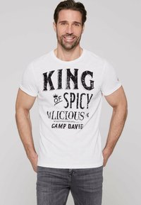 Weißes Baumwoll-T-Shirt mit schwarzem Drucktext: "KING of SPICY" und "CAMP DAVID." Rundhalsausschnitt, kurze Ärmel, lässige Passform. Graue Jeans getragen.