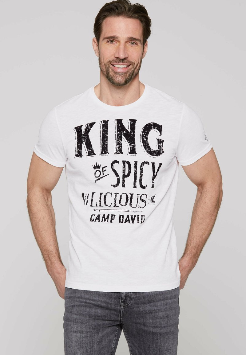 Weißes Baumwoll-T-Shirt mit schwarzem Drucktext: "KING of SPICY" und "CAMP DAVID." Rundhalsausschnitt, kurze Ärmel, lässige Passform. Graue Jeans getragen.