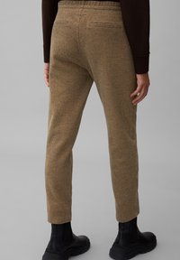 Tapered broeken in een gestructureerde bruine stof met een subtiel ruitpatroon, voorzien van een elastische tailleband met trekkoord en twee achterzakken. Zwarte enkellaarzen afgebeeld.
