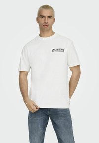 Maglietta in cotone bianco con scollo rotondo e maniche corte, caratterizzata da una stampa nera del logo sul lato sinistro del petto. Indossata con jeans in denim blu.