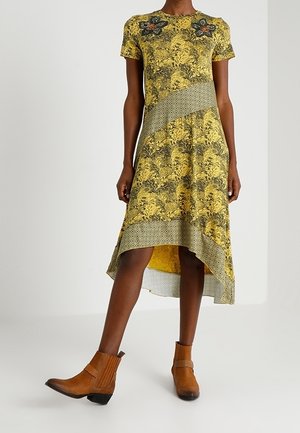 Kobieta w żółtej sukience o wzorze, krótszej z przodu i dłuższej z tyłu, z kwiatowymi detalami oraz brązowymi botkami do kostki, stoi z jedną nogą lekko uniesioną.