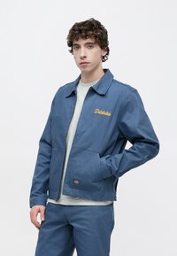 Μπλε καμβά τζάκετ με φερμουάρ στο μπροστινό μέρος, μυτερό γιακά και κεντημένο το λογότυπο "Dickies" με κίτρινο χρώμα. Διαθέτει πλαϊνές τσέπες και ομαλή υφή.