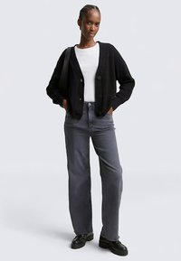 Cardigan noir côtelé avec fermeture à boutons, porté sur un haut blanc, associé à un jean gris et des chaussures noires, avec une coupe décontractée.