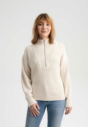 Beige knit pullover with high collar, half-zip closure, and textured pattern; features wide sleeves and ribbed hem, worn over light jeans.
