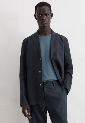Blazer en lin bleu foncé avec boutonnage simple, revers à cran, deux poches avant et trois boutons. Porté sur un tee-shirt bleu clair à col rond.