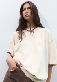 Beige übergroßes T-Shirt mit kurzen Ärmeln, kombiniert mit braunen Cordhosen. Eine Kettenhalskette mit einem orangefarbenen Akzent ist sichtbar.