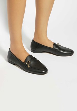 Mocassins en cuir noir avec détail de boucle dorée portés aux pieds, présentés sur un fond blanc.
