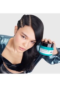 Turkos färgad burk märkt "Scalp Advanced" med ett svart lock, hålls i en hand. Modellen har släta mörka hår och bär en glänsande svart topp, tillsammans med en blänker blå jacka.