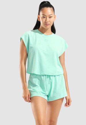 Kvinde med langt mørkt hår samlet i en hestehale iført mintgrøn ærmeløs top og matchende shorts mod en ensfarvet lys baggrund.