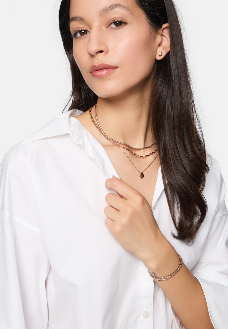 Tre lagdelte halskæder i rosaguld, en lille vedhæng med oval form og et matchende armbånd. Modellen har en hvid skjorte med knapper på.