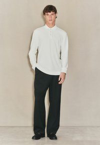 Ilgų rankovių balta polo marškinėliai su apykakle ir trimis mygtukais, derinami su juodais plačiais kelnais ir juodais batais. Paprastas, švarus dizainas.