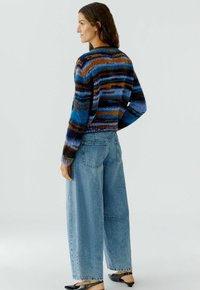 Strickpullover mit mehrfarbigen Streifen in Blau, Braun und Schwarz; kombiniert mit lockeren hellblauen Jeans und schwarzen Ballerinas.