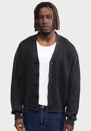 Ung mand med kort fletning, iført en sort ribbet cardigan over en hvid skjorte og blå jeans, stående mod en ensfarvet baggrund.