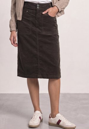 Femme portant une jupe en velours côtelé marron foncé longueur genou avec poches avant, une veste beige et des baskets blanches avec des rayures bordeaux.