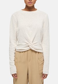 Haut crème à manches longues légèrement texturé avec un détail torsadé à l'avant, porté avec un pantalon beige doté de poches latérales. Tissu doux, coupe décontractée.