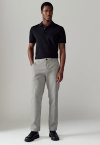 Polo noir à manches courtes avec un col à trois boutons associé à un pantalon gris clair, comportant une coupe droite et des poches avant, porté avec des chaussures noires.