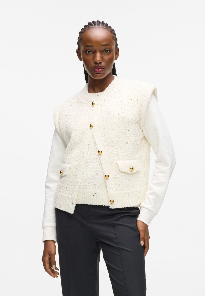 Krēma tekstūras kardigāns ar zelta pogām, ar divām priekšējām kabatām. Uzvilkts virs baltas garām piedurknēm blūzes, saskaņots ar tumšajiem biksēm.