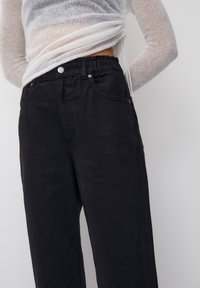 Pantaloni neri, larghi e con elastico in vita, dotati di tasche frontali e dettagli di cucitura discreti. Il tessuto sembra morbido e leggero.