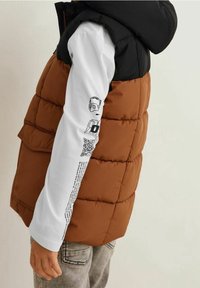 Brązowa kamizelka puchowa z czarnym kapturem, z pikowanym wzorem i zamykana na kieszonkę z przodu. Biała koszula z długim rękawem z nadrukowanymi grafikami i tekstem.