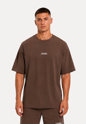 Un giovane uomo è in piedi, rivolto in avanti, indossa una t-shirt marrone larga e pantaloncini abbinati con piccoli loghi bianchi su entrambi i capi.