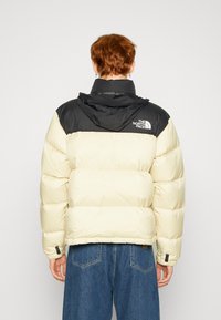 Crème en zwarte pufferjas met een capuchon, voorzien van quilted segmenten en een merklogo op de achterkant. Soepele textuur en een relaxte pasvorm.