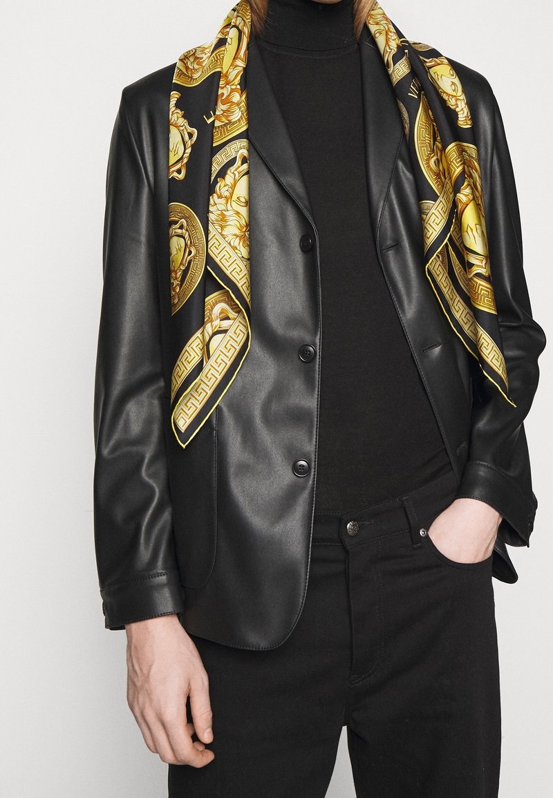 Personne portant un blazer en cuir noir, un col roulé noir, un pantalon noir et une écharpe noire et dorée à motifs drapée sur les épaules.