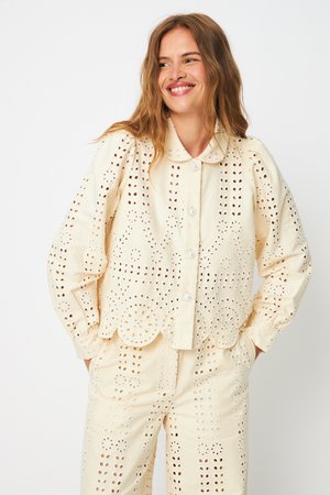 Pikkade juustega naine naeratab, kandes kreemjat õmblustega pluusi sakilise äärega ja sobivaid pükse, käed taskutes.