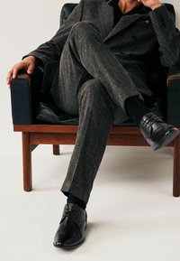 Zapatos de vestir de cuero negro con acabado suave, diseño de cordones y punta redondeada, llevados con un traje gris texturizado y calcetines oscuros.
