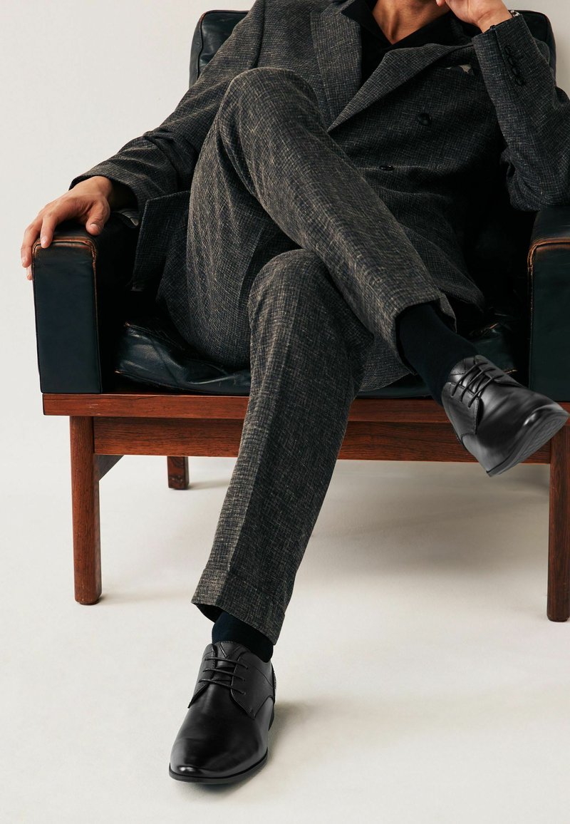 Zapatos de vestir de cuero negro con acabado suave, diseño de cordones y punta redondeada, llevados con un traje gris texturizado y calcetines oscuros.