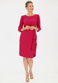 Jasnoróżowa sukienka z rękawami o długości trzech ćwierci, dopasowana sylwetka oraz falbana po boku. Gładki materiał o subtelnym połysku.