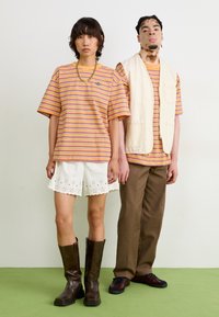 Randig orange och lila t-shirt matchad med vita shorts med hålmockad kant; bruna knähöga stövlar och avslappnade bruna byxor.