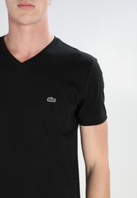 Černé bavlněné tričko s výstřihem do V. Na levé straně hrudi se nachází malý barevný logo krokodýla Lacoste. Krátké rukávy.