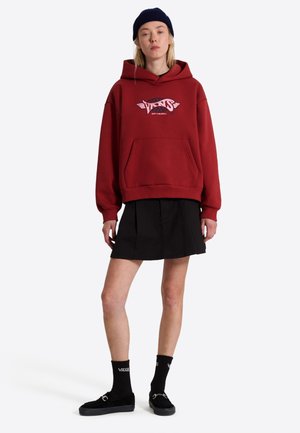 Ung person iført en rød Vans hættetrøje, sort nederdel, sorte sokker med Vans-logo, sorte sko og en sort beanie, stående mod en hvid baggrund.