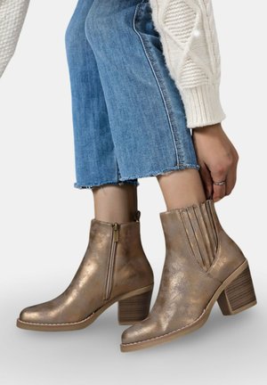 Bottines en bronze avec une finition métallique lisse, fermeture éclair, accents nervurés et un talon épais. Portées avec un jean bleu court.