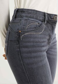 Vaqueros de mezclilla gris oscuro con una cintura alta, costuras en contraste y un pequeño diseño de bolsillo decorativo. Acentos en tono cobre.