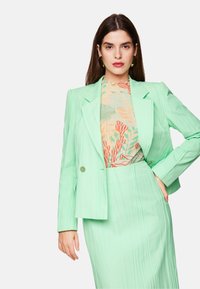 Mint green tailored blazer with a structured design, paired with a patterned blouse featuring floral elements in orange and green tones.