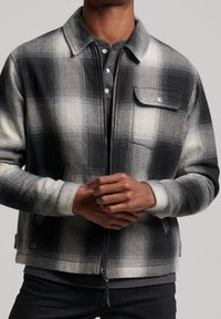 Schwarz-graue karierten Jacke mit durchgehendem Reißverschluss, mit Knöpfen versehener Brusttasche und gerippten Bündchen. Hergestellt aus weichem Stoff, der eine strukturierte Form bietet.