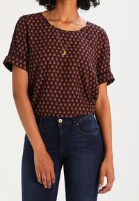 Kvinna som bär en mönstrad mörkbrun blus med korta ärmar och uppvikta ärmar, samt mörkblå jeans, accessoriserad med ett halsband med en halvmåne.