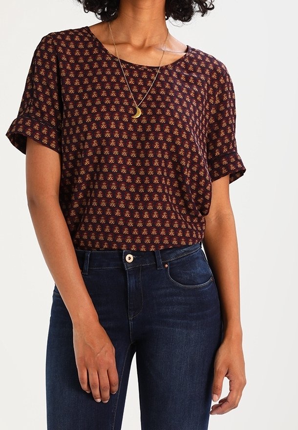 Kvinna som bär en mönstrad mörkbrun blus med korta ärmar och uppvikta ärmar, samt mörkblå jeans, accessoriserad med ett halsband med en halvmåne.