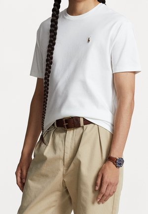 White cotton t-shirt with short sleeves, featuring a small embroidered logo on the left chest, paired with beige pleated pants and a brown belt.