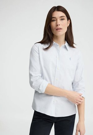 Vrouw met stijlbruin haar die een wit en lichtblauw gestreept overhemd met opgerolde mouwen en een zwarte broek draagt.