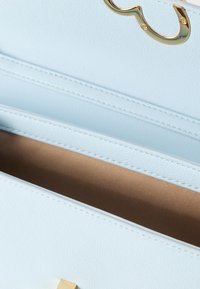 Τσάντα χειρός από ανοιχτό μπλε δέρμα με ομαλή υφή, χρυσά μεταλλικά στοιχεία, λεπτομερείς ραφές και καφέ υφασμάτινη επένδυση που διακρίνεται κάτω από το ανοιχτό καπάκι.