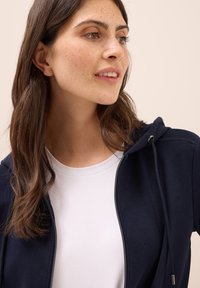 Haut à capuche zippé bleu marine avec cordon de serrage, fabriqué en tissu texturé, porté sur un t-shirt blanc à col rond uni. Gros plan du visage et du cou d'une personne.