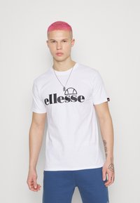 Hvit bomull t-skjorte med en stor svart "ellesse" logo på forsiden. Korte ermer, rund hals og en liten logo aksent på ermene.