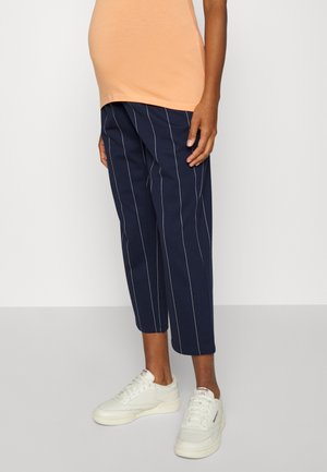 Osoba ubrana w granatowe spodnie 7/8 z białymi pionowymi paskami, brzoskwiniowy top i białe trampki, stojąca na jasnoszarej podłodze.