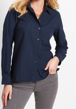 Femme portant une chemise bleu marine à boutons, un pantalon gris, un collier en or et des bagues, une main dans la poche et l'autre détendue.
