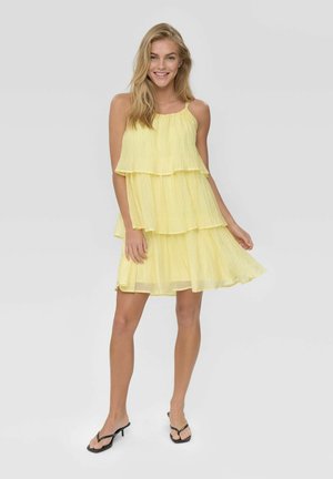 Hymyilevä nainen, joka pukeutunut hihattomaan keltaiseen kerrokselliseen mekkoon ja mustiin korkokenkiin, seisoo yksinkertaisen vaalean taustan edessä.