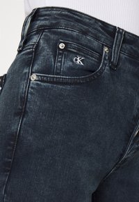 Mörka denimjeans med en tajt passform, subtila blekningar, silverfärgade nitar och en vit broderad logotyp på framfickan.