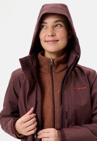 Bordeaux waterdichte jas met capuchon, voorzien van ritszakken en een zachte bruin fleecelaag aan de binnenkant. Vaude-logo in oranje op de borst.