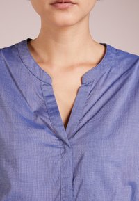 Blusa de algodón azul con escote en V y mangas cortas. Textura suave con un brillo sutil; cuenta con un cuello doblado y tapeta frontal.
